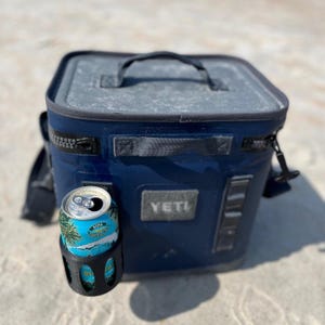 Op de afbeelding: Een marineblauwe YETI-koeler met een zwarte handgreep en ritssluiting, en een zwarte bekerhouder. Een blikje Kona Brewing Co.-bier zit in de bekerhouder. De koeler staat op een zanderige ondergrond.