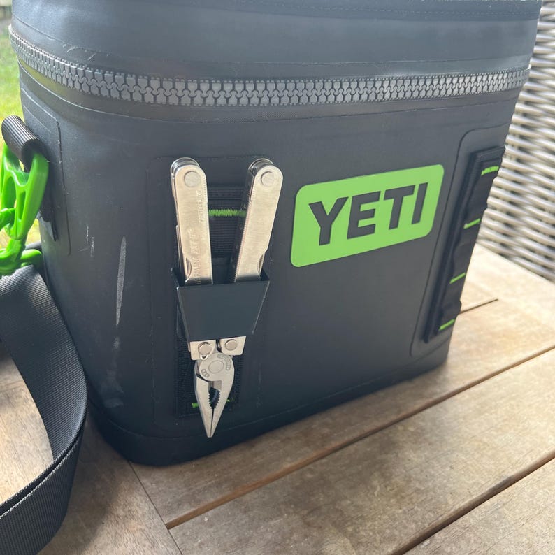 May include: Black insulated cooler bag with a green and black strap and zipper. The bag has a green "YETI" logo and a black holder with a multi-tool. The multi-tool is silver and has pliers.