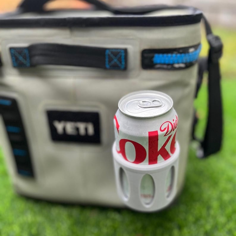 Op de afbeelding: Een wit en rood Diet Coke-blikje in een witte blikhouder, voor een grijze YETI-koeler. De koeler heeft zwarte en blauwe accenten, een zwarte handgreep en een zwarte schouderriem. De achtergrond is groen gras.