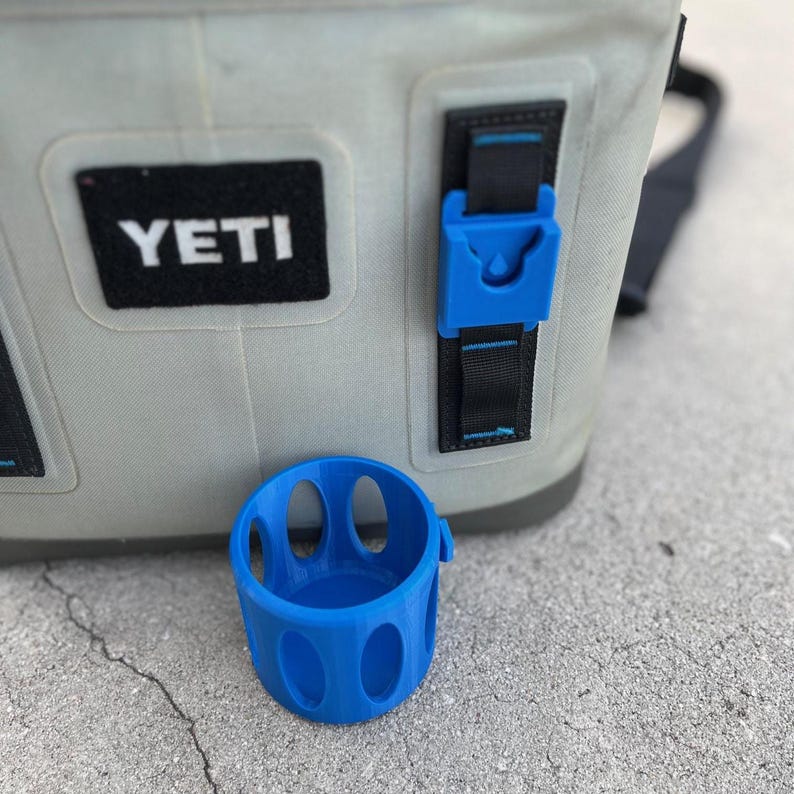Op de afbeelding: Een blauwe bekerhouder met ovale uitsparingen staat voor een grijze koeler. De koeler heeft een zwart "YETI"-patch en een blauwe gesp. De bekerhouder is ontworpen om aan de riem van de koeler te bevestigen.