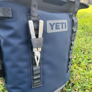 May include: A navy blue YETI cooler with a black strap holding a silver multi-tool. The multi-tool is in a black holder attached to the cooler. The YETI logo is visible on the cooler.