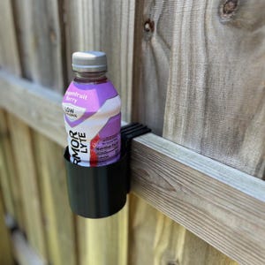 May include: A black cup holder attached to a wooden fence holds a bottle of Armor Lyte drink. The bottle is purple and white with the words "Dragonfruit Berry" and "LOW CALORIE" visible. The fence is made of weathered wood.