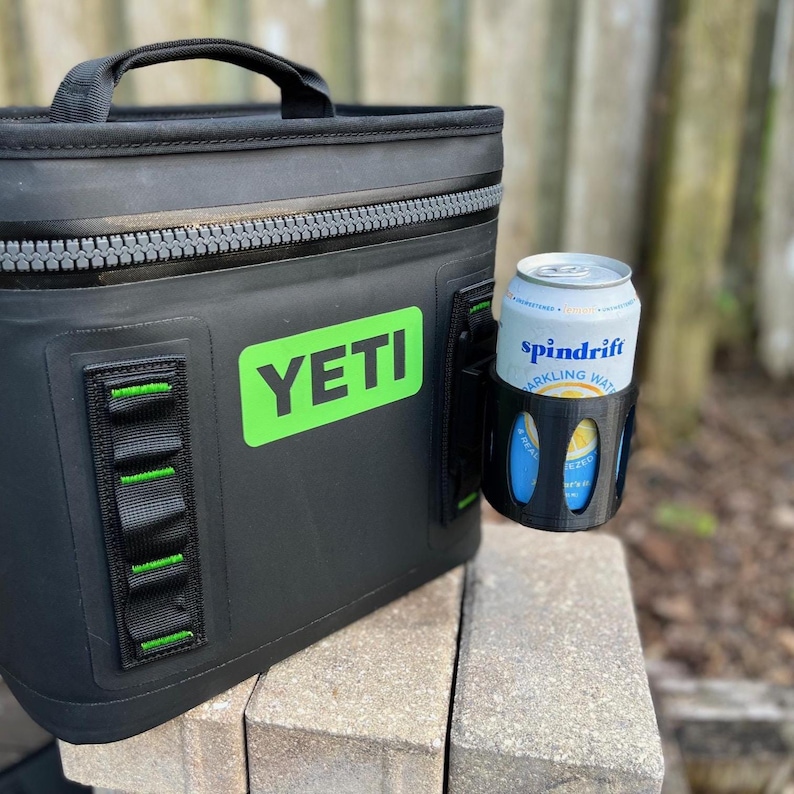 Op de afbeelding: Zwarte Yeti-koeler met een groen logo en een zwarte bekerhouder. Een blikje Spindrift bruisend water zit in de bekerhouder. De koeler heeft een handvat en een ritssluiting.