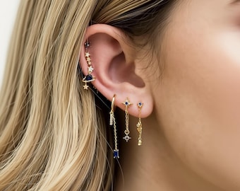 Op de afbeelding: Een verzameling gouden oorbellen met hemelse ontwerpen. De oorbellen hebben sterren, een planeet en blauwe edelstenen. Eén oorbel is een hoepel met een bungelende ketting en een blauwe rechthoekige steen. Andere oorbellen zijn studs en bungelende oorbellen.