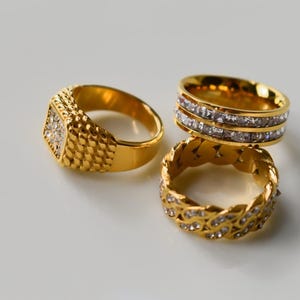 May include: Three gold rings are shown on a white surface. One ring features a square face with a diamond pattern, another has two rows of clear stones, and the third has a braided design with clear stones.