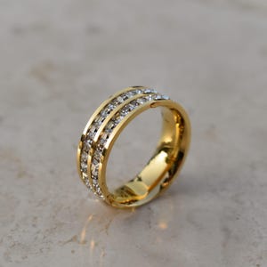 May include: A gold-colored ring with three rows of clear, sparkling gemstones. The ring is set against a neutral background, highlighting its polished, reflective surface. This piece of jewelry is a classic design.