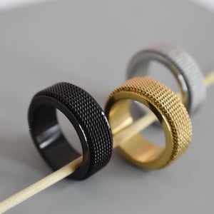 May include: Three metal rings in black, gold, and silver, each with a textured surface. The rings are displayed on a light gray surface, with a wooden stick running through each ring. The rings are likely fashion accessories.