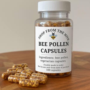 May include: A clear plastic bottle with a white lid and a label that reads "BEE POLLEN CAPSULES." The label also includes the text "SHOP FROM THE HIVE" and a bee graphic. Several capsules are scattered in front of the bottle.
