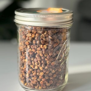 May include: A clear glass jar filled with brown granules, sealed with a silver lid. The jar is labeled with the word "Ball" and is sitting on a white surface. The contents appear to be bee pollen.