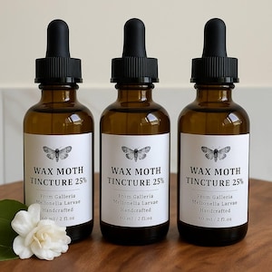 Peut inclure: Trois flacons en verre ambré avec des compte-gouttes noirs, chacun étiqueté "WAX MOTH TINCTURE 25%" avec une illustration de papillon. Les étiquettes indiquent également "From Galleria Mellonella Larvae Handcrafted 60 ml / 2 fl.oz". Une fleur blanche est au premier plan.