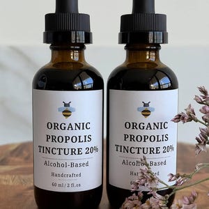Puede incluir: Dos frascos de vidrio ámbar con tapas cuentagotas negras, cada uno con una etiqueta blanca con un icono de abeja y el texto Organic Propolis Tincture 20%, Alcohol-Based, Handcrafted, 60 ml / 2 fl. oz. Los frascos contienen un líquido oscuro.