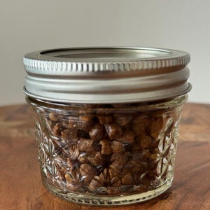 May include: A small, clear glass jar with a silver lid, filled with brown, granular material. The jar has a decorative, embossed pattern. The contents appear to be small, irregularly shaped pieces. The jar sits on a wooden surface.