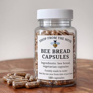 以下が含まれることがあります： 白い蓋が付いた透明なガラス瓶に、ミツバチのパンカプセルが詰まっています。「Shop From The Hive」と「Bee Bread Capsules」と書かれたラベルが付いています。ラベルには成分とカプセルの数も記載されています。カプセルは茶色です。