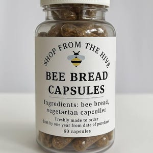 Pode incluir: Um frasco de plástico transparente com tampa branca de rosca contendo cápsulas castanhas. O rótulo branco apresenta um ícone de abelha e o texto: SHOP FROM THE HIVE, BEE BREAD CAPSULES, Ingredients: bee bread, vegetarian capculler, 60 capsules.