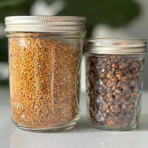 May include: Two clear glass mason jars with silver metal lids. The larger jar on the left is filled with golden-yellow bee pollen granules. The smaller jar on the right contains dark brown, textured propolis pieces. Both jars have a textured surface.