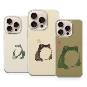 May include: Three phone cases featuring frog illustrations. The cases have a clear border and a beige, cream, and olive green background. The frog designs are in shades of green and brown, with Japanese-style art.