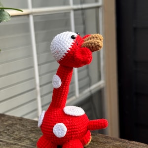 Könnte beinhalten: Ein handgefertigtes Häkeltier in Form einer Giraffe. Die Giraffe ist hauptsächlich rot mit weißen Flecken und einem weißen Kopf. Schnauze und Füße sind braun. Das Spielzeug steht auf einer Holzoberfläche.