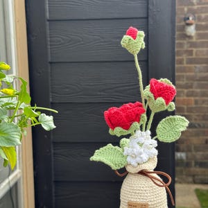 Könnte beinhalten: Ein handgefertigtes Häkelblumenarrangement mit roten Rosen, grünen Blättern und weißen Blüten in einer beigen Vase. Die Vase hat eine braune Lederschleife und ein Etikett mit der Aufschrift "keep your smile".