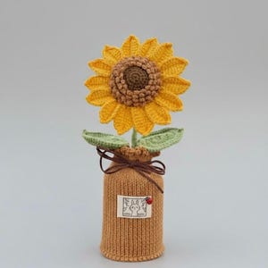 Peut inclure: Un tournesol crocheté à la main, avec des pétales jaunes et un centre marron, placé dans un vase en tricot marron. Le vase porte une petite étiquette avec un motif de lapin et une coccinelle. La marque "AUSASI HANDICRAFTS" est visible.
