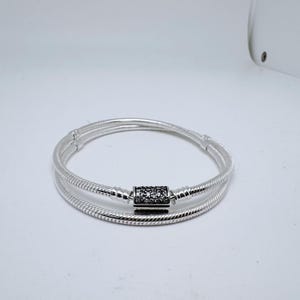 May include: A silver bracelet with a snake chain design. The bracelet has a rectangular clasp set with small, dark stones. The item is shown against a white backdrop.