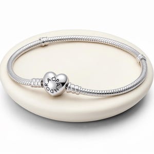 Puede incluir: Una pulsera plateada con un cierre en forma de corazón. El cierre presenta la palabra "PANDORA" y la pulsera tiene un diseño de cadena de serpiente. La pulsera se muestra sobre una superficie blanca de forma ovalada.