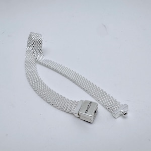 Könnte beinhalten: Ein silbernes Armband mit einem Mesh-Muster und einem rechteckigen Verschluss. Der Verschluss trägt die Aufschrift "Pandora". Das Armband ist geöffnet und liegt auf einer weißen Oberfläche. Das Mesh-Muster ist sichtbar.
