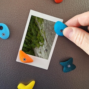 Może przedstawiać: Zdjęcie w stylu polaroid przedstawiające wspinacza górskiego na ścianie skalnej, otoczonego kolorowymi chwytami wspinaczkowymi. Chwyty mają różne kształty i kolory, w tym niebieski, pomarańczowy i żółty. Obraz jest na brązowej powierzchni.