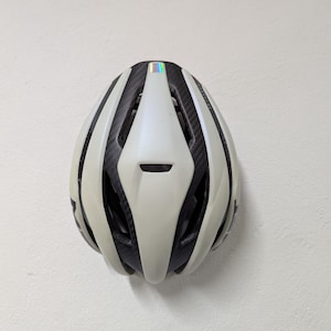 May include: A white and black bicycle helmet with a streamlined design. The helmet has a glossy finish with black vents and a carbon fiber weave pattern. A small, iridescent sticker is on top.