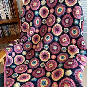May include: A crocheted blanket in shades of blue, pink, yellow, and purple, draped over a chair. The blanket's design features numerous circular patterns. A decorative and cozy home accessory.