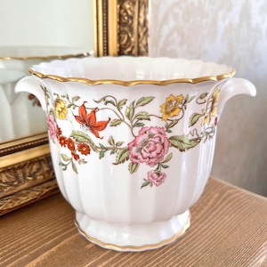 May include: A white porcelain planter with a fluted body and two side handles. The exterior features a colourful floral pattern with pink, orange, and yellow flowers and green leaves. The rim and base are finished with a thin gold trim.