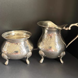 May include: A silver-toned metal sugar bowl and creamer set. Both pieces feature an engraved floral pattern, a rounded body, and three feet with a shell-like design. The creamer includes a curved handle and a pouring spout for liquid service.