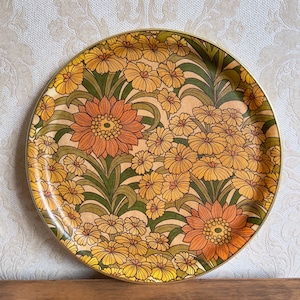 May include: A round serving tray featuring a dense floral pattern with large orange and yellow flowers interspersed with smaller yellow blossoms and green foliage on a light tan background, finished with a thin metallic gold-toned rim.