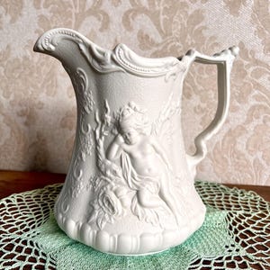 May include: A white ceramic pitcher featuring an embossed relief of a cherub resting among floral vines. The vessel has a textured surface, a scalloped base, and an ornate handle. The rim is curved with decorative molding along the top edge.