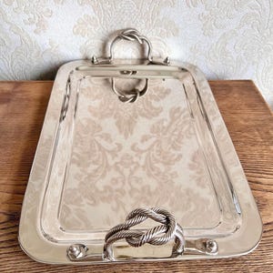 May include: A rectangular silver-toned serving tray with a polished, reflective surface. The tray features two handles, each designed in the shape of a decorative nautical knot, securely attached to the short ends with small metal rivets.