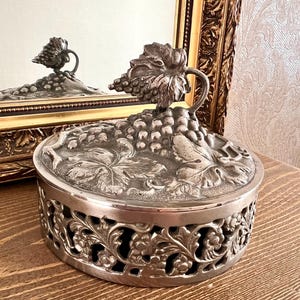 May include: A round silver-toned metal container with a lid featuring a raised grape cluster and leaf design. The sides of the container display an intricate openwork pattern of vines and leaves, creating a decorative, textured metallic finish.