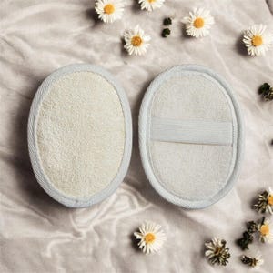 May include: Two oval-shaped, off-white bath loofahs with a light blue trim. One loofah has a hand strap. The loofahs are on a soft, textured surface, with scattered white daisies.