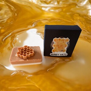 Könnte beinhalten: Ein braunes und weißes Seifenstück mit einem Wabenmuster, neben einer schwarzen Schachtel mit einer Waben-Grafik und dem Text "HONEY OATS". Der Hintergrund ist eine goldene, flüssige Textur.
