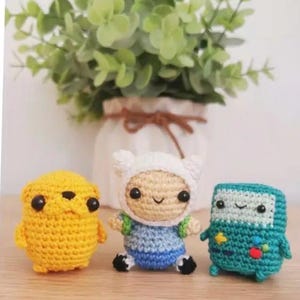 May include: Three handmade crochet figures: a yellow dog-like character, a figure in a white hat and blue outfit, and a teal robot. The figures are on a wooden surface, with a vase of greenery in the background.