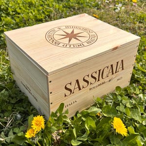 Könnte beinhalten: Eine hellfarbene Holzkiste mit dem Wort "SASSICAIA" prominent dargestellt. Die Kiste trägt auch den Text "BOLGHERI 2023" und "ITALIA". Die Oberseite der Kiste hat ein Kompassrosen-Design. Die Kiste ist von grünem Gras und gelben Blumen umgeben.