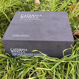 Puede incluir: Una caja negra con el texto "CATERINA RIVELLA ALTA LANGA ROSATO RISERVA" y "MARCALBERTO METODOCLASSICO" impreso. La caja está sobre hierba verde. La caja contiene 3 botellas de vino.