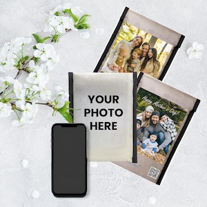 Könnte beinhalten: Ein Smartphone liegt neben einer personalisierten Fototasche mit dem Text "YOUR PHOTO HERE". Zwei weitere Taschen zeigen Familienfotos, eine davon mit dem Text "Happy Mother's Day!". Weiße Blumen und ein heller Hintergrund vervollständigen die Szene.