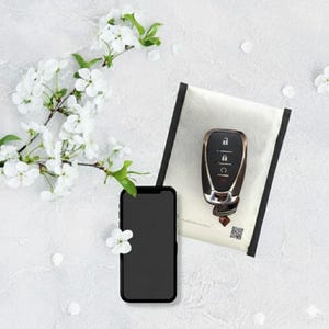 Könnte beinhalten: Ein schwarzes Smartphone und ein Autoschlüsselanhänger liegen auf einer weißen Oberfläche, neben einem weißen Beutel mit schwarzem Rand. Weiße Kirschblüten und Blütenblätter sind um die Objekte verstreut.