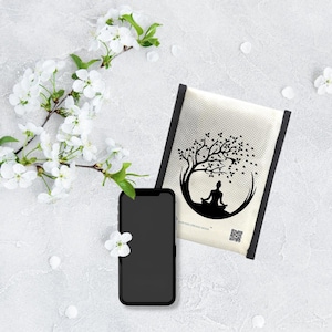 May include: A black smartphone with a blank screen rests near a cream-colored meditation pillow. The pillow features a black tree and figure design. White flowers and petals are scattered around the objects.