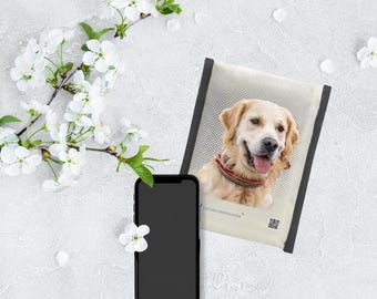 May include: A smartphone with a blank black screen rests near a cream-colored pet portrait. The portrait features a Golden Retriever with a red and black collar. White flowers and petals are scattered around the objects.