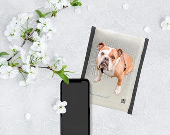 May include: A tan and white bulldog rests on a light beige pet bed with a black border. A black smartphone and a flowering branch are also in the image. The pet bed has the text "SAFE KIDS STRONG HOME".