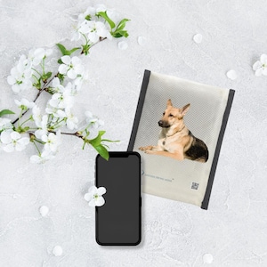 Könnte beinhalten: Ein Smartphone mit schwarzem Bildschirm liegt neben einem Hundebett mit einem Deutschen Schäferhund. Das Hundebett hat einen grauen Rand und eine helle Oberfläche mit dem Text "SAFE FOR STRONG HOME". Weiße Blüten und Blütenblätter sind verstreut.