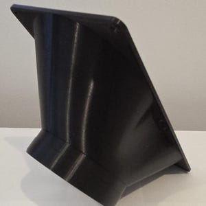 Puede incluir: Un objeto negro y angular con una parte superior trapezoidal y una base cilíndrica. El objeto tiene una superficie lisa y reflectante y parece estar hecho de plástico. El diseño presenta una abertura acampanada en la parte superior y una abertura más pequeña en la parte inferior.