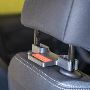 May include: A dark grey plastic car headrest mount featuring two circular clamps that attach to the metal headrest posts. The mount includes a rectangular orange accent piece with diagonal line detailing on its front-facing surface.