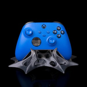 Peut inclure: Une manette Xbox bleue est posée sur un support noir au design stylisé. La manette affiche le logo Xbox et les boutons de commande. Le support a une forme angulaire, et le fond est noir.