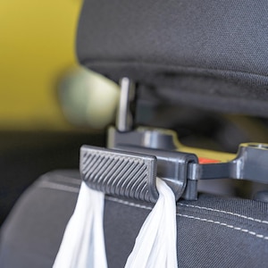May include: A dark gray plastic car headrest hook featuring a textured, ribbed surface on the front edge. The hook is securely attached to the metal posts of a black fabric car seat headrest, with the white handles of a plastic bag hanging from it.